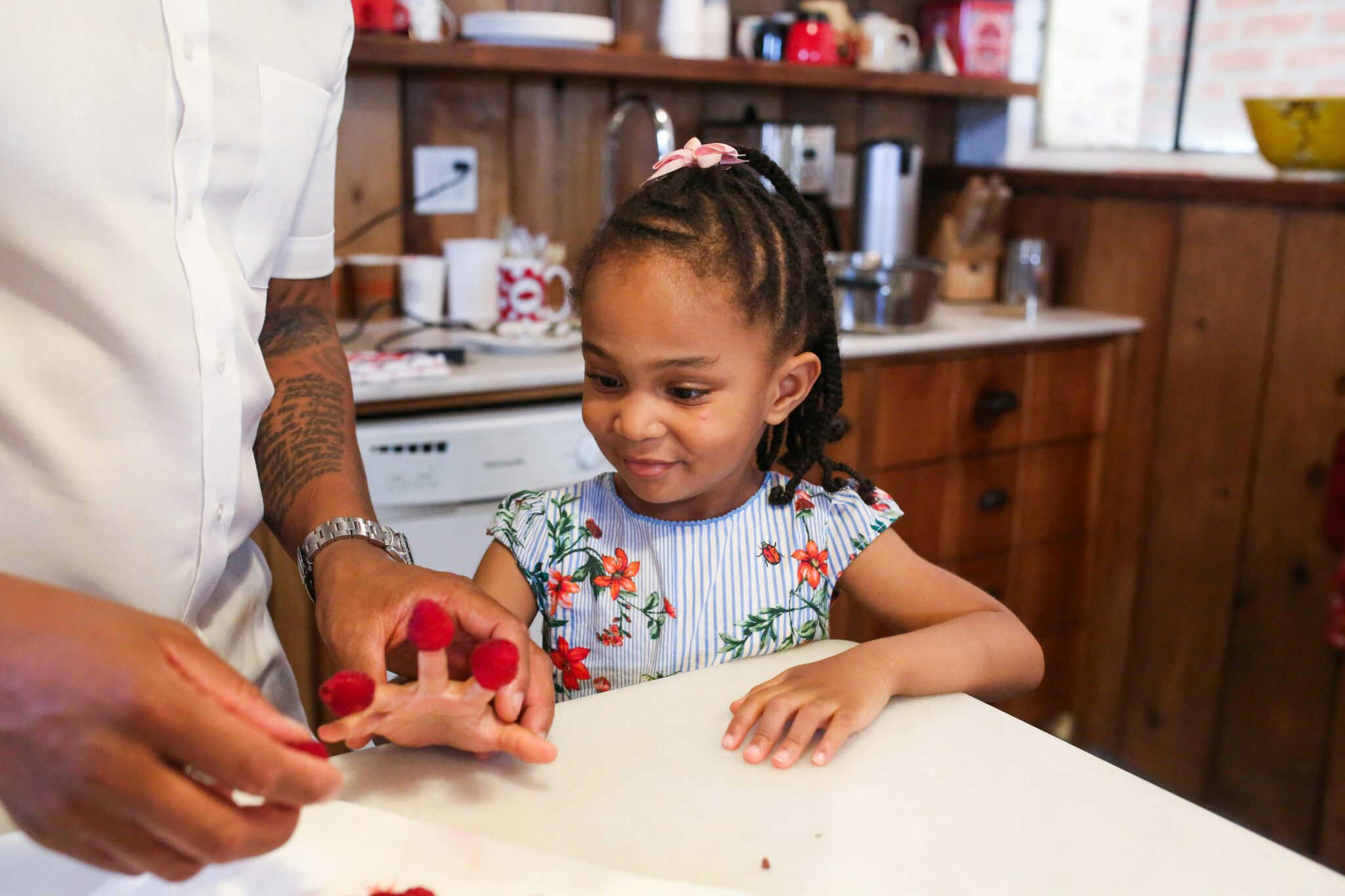 Father and daughter in the kitchen, with the girl placing raspberries on her fingers, illustrating cooking techniques, food preparation, and kitchen basics. This scene highlights the importance of culinary education, knife safety, and engaging children in learning knife skills and cooking techniques for beginners.