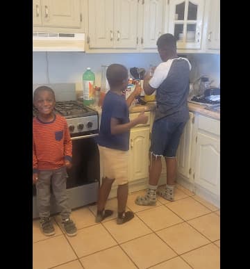 Smiling Black father and son pouring fresh green juice into a glass in a bright kitchen. Healthy homemade juicing, plant-based nutrition, and family wellness. Fresh fruits and vegetables in the background.
