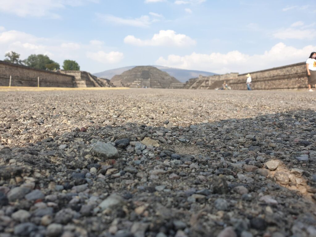 Ground-level view of the Avenue of the Dead leading toward a famous pyramid at Teotihuacán in Mexico, with stone ruins, distant mountains, and a bright blue sky.