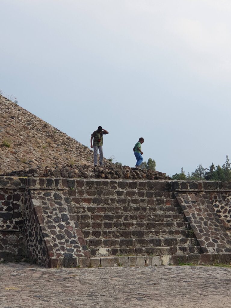 Father and son exploring the stone steps and lower platform of an ancient pyramid in Mexico, with a wide open sky above.