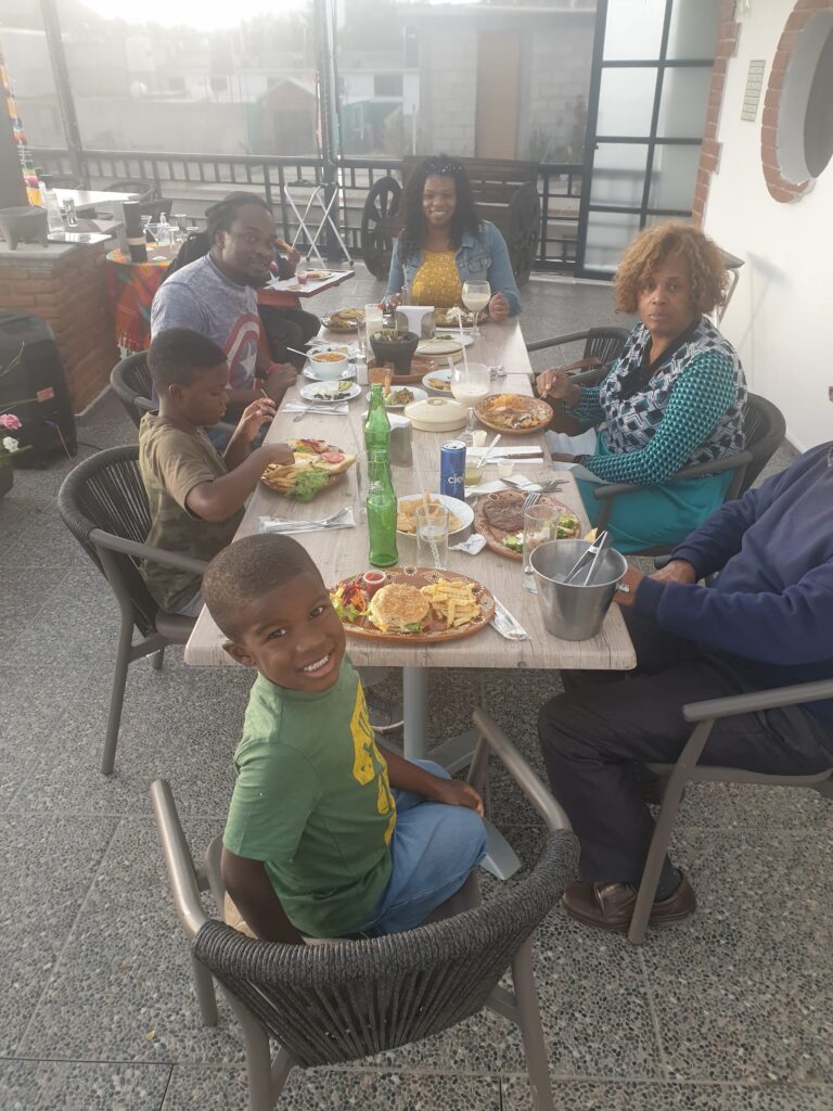 Family enjoying a meal together at an outdoor restaurant in Mexico, with plates of food on the table and a young boy smiling at the camera in the foreground.