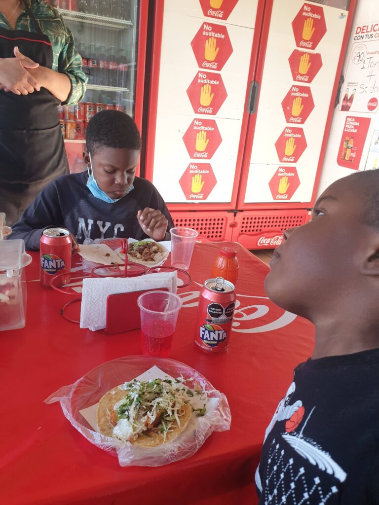 Two boys sit at a red table in a casual taco shop in Mexico, eating tacos and drinking soda, with a Coca-Cola fridge and a server in the background.